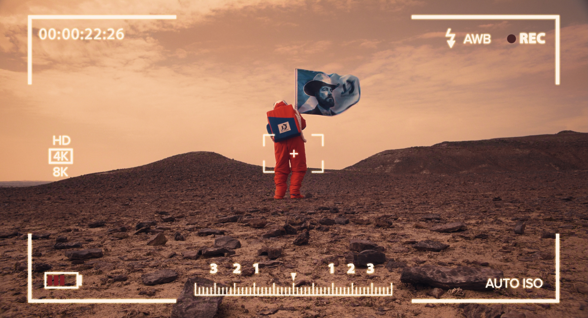 A figure in an orange suit holding a flag stands on rocky terrain under a faded sky.