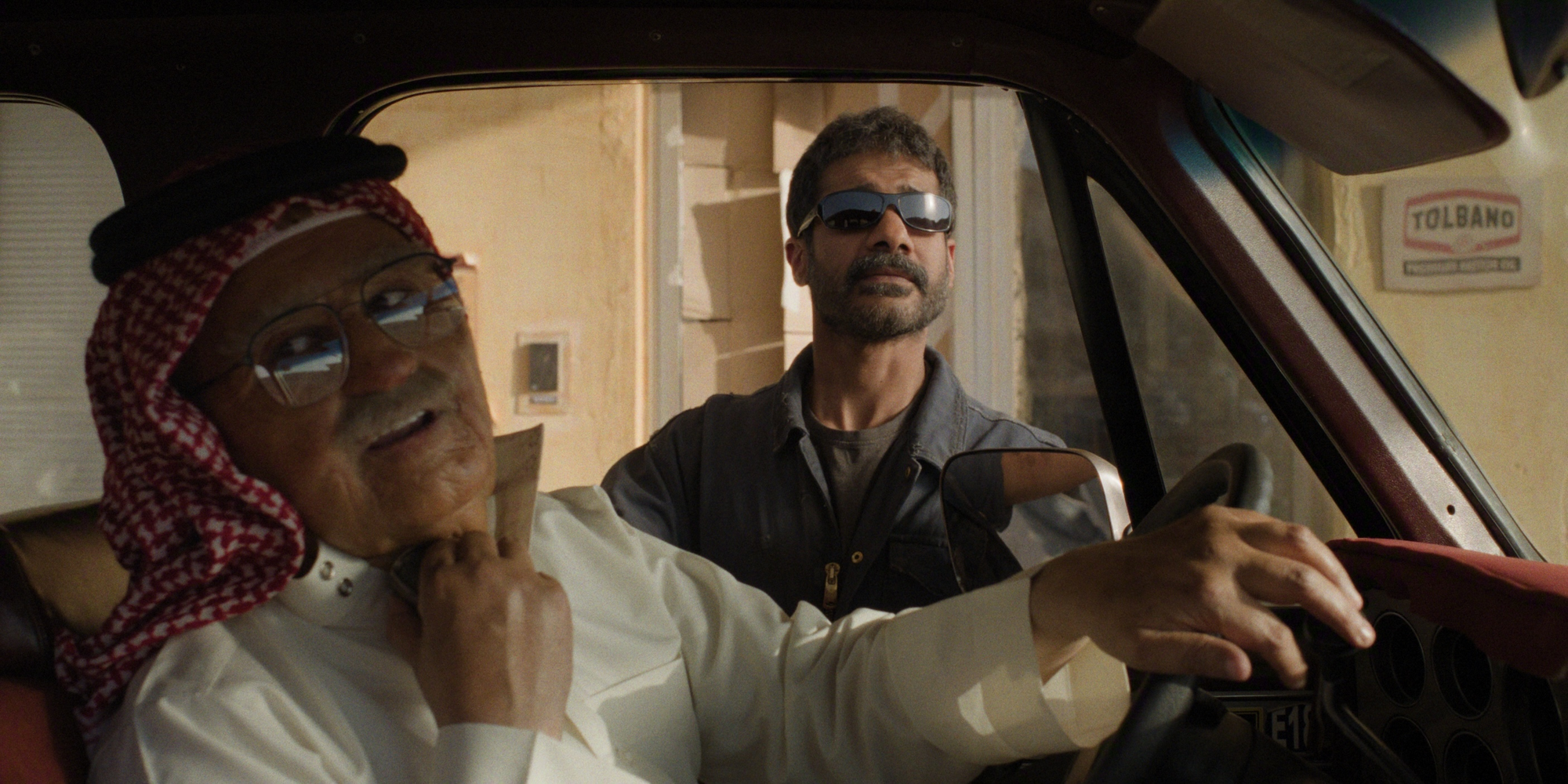 Two men in a vehicle, one in traditional attire and the other in sunglasses, engaged in conversation.