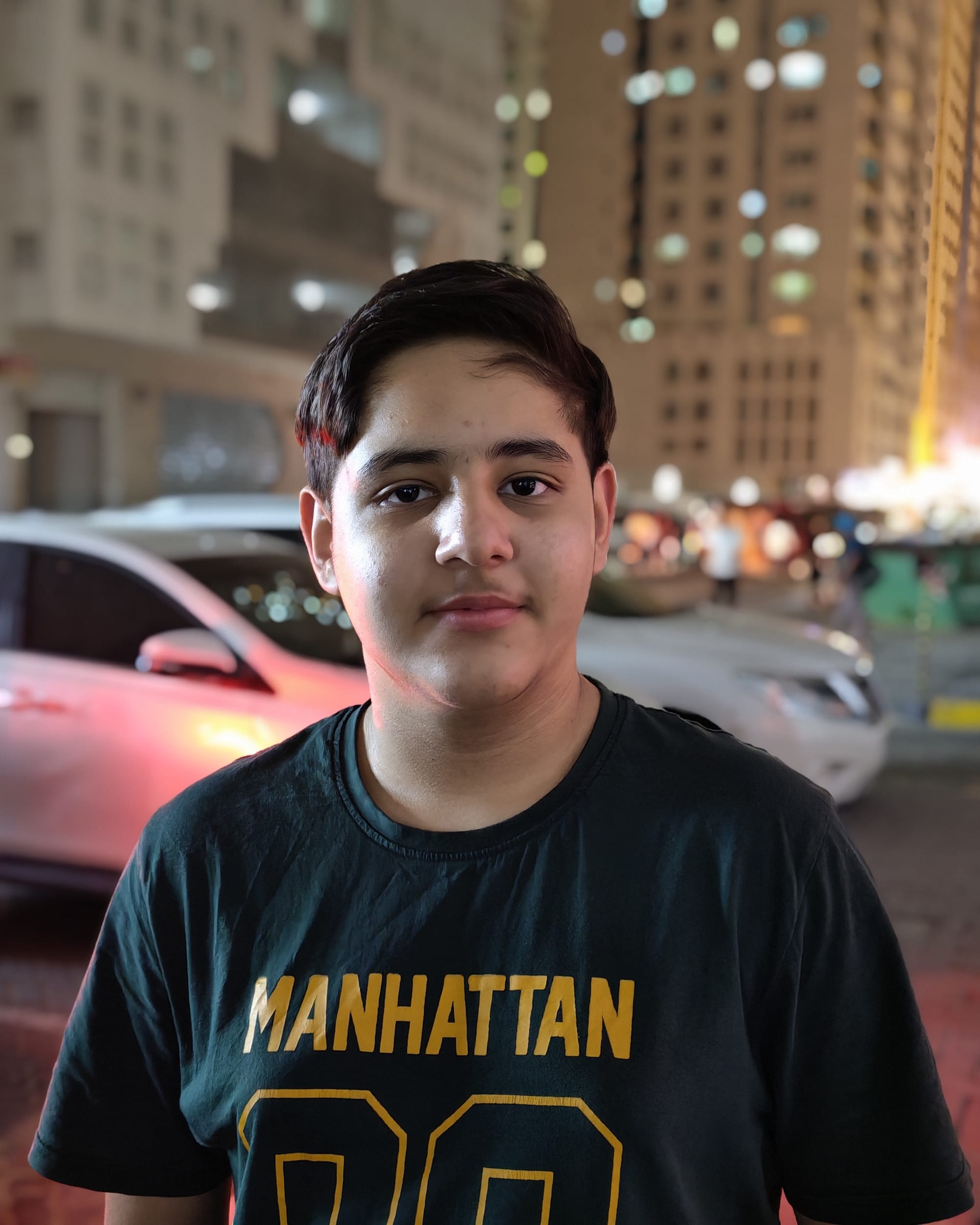 Honor 600 night portrait camera sample showing a subject against bokeh-blurred Sharjah apartment buildings and parked cars