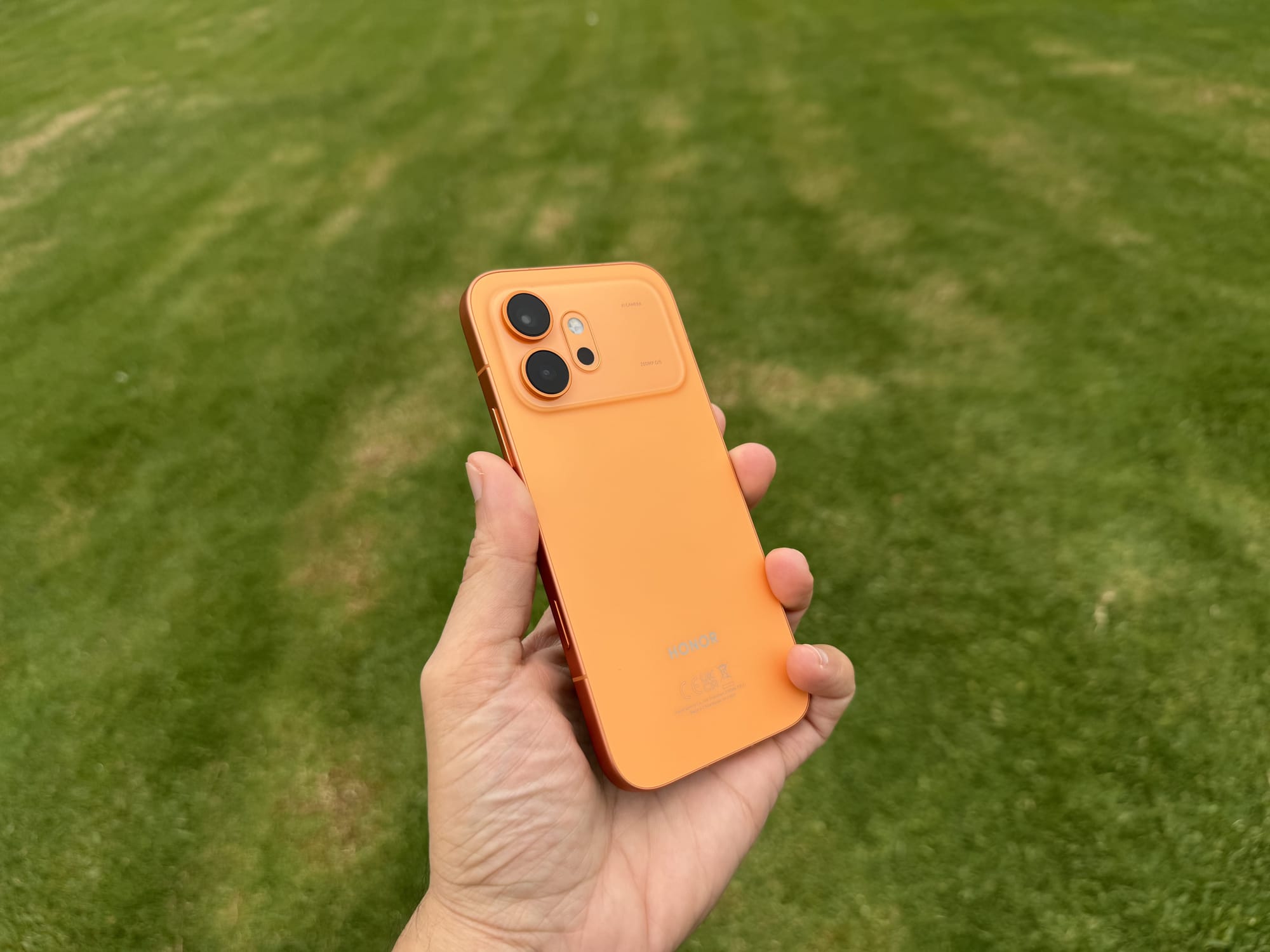 Rear three-quarter view of the Honor 600 in orange held over grass, showing the slim profile and camera island