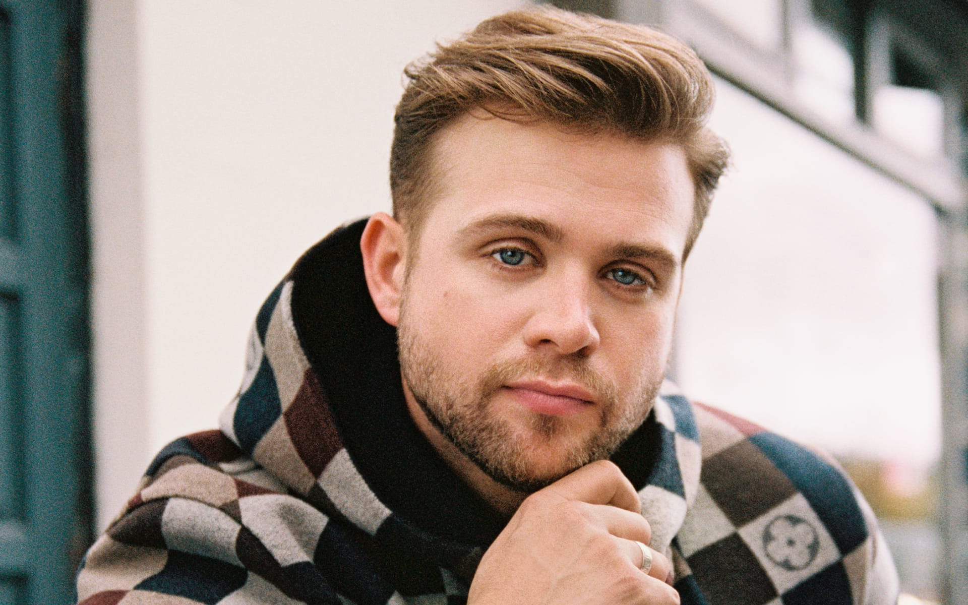 Leo Woodall poses thoughtfully in a patterned hoodie with a blurred background.
