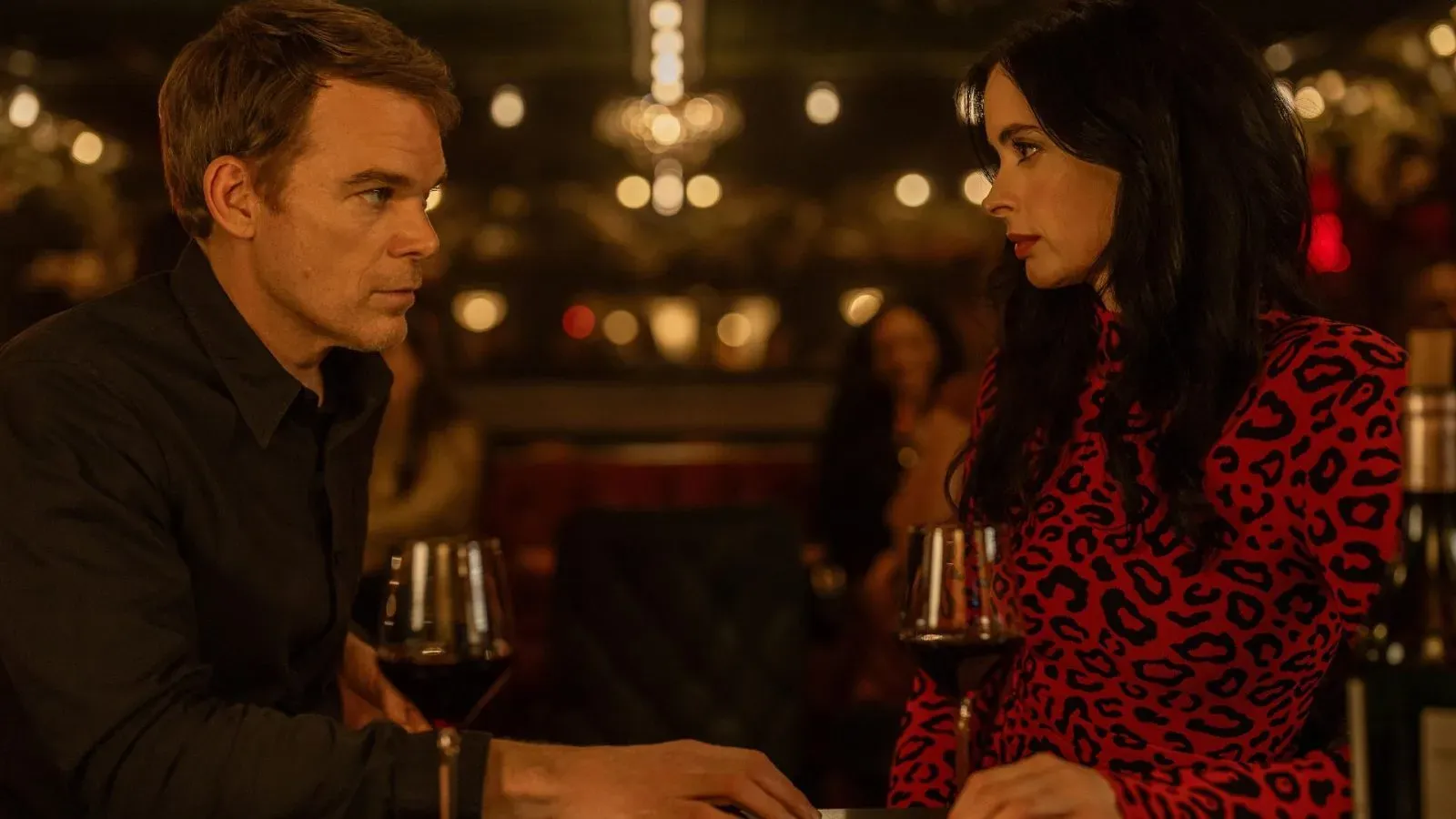 Michael C. Hall and a woman in a vibrant leopard print dress share an intense conversation at a bar.