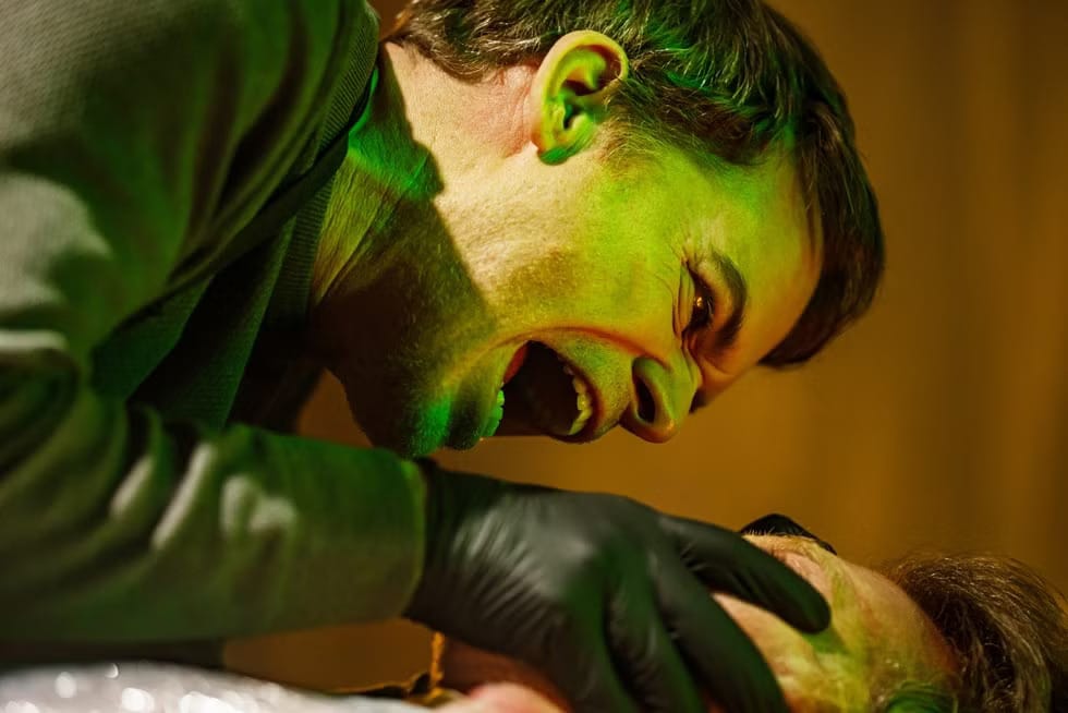 A frantic scene with a man shouting while gripping another man's face, illuminated with green light.