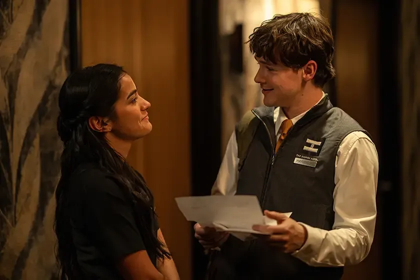 A woman smiles at a man in a vest as he hands her a document in a sleek hallway.