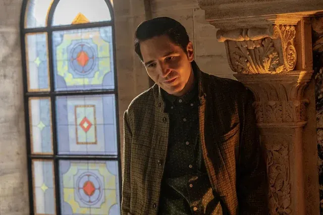 A man leans against a stone pillar, thoughtfully gazing into the distance with stained glass in the background.