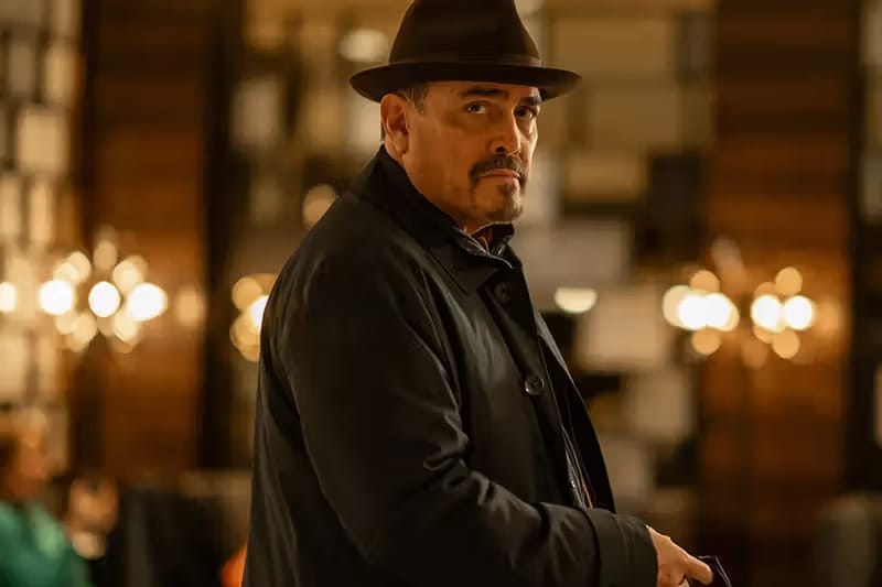 A man in a hat and coat stands in a classy environment, looking back with a serious expression.