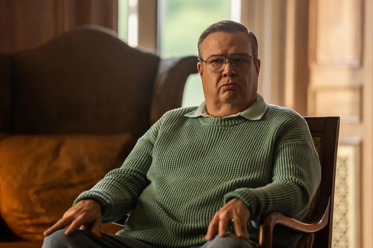 A seated man in a green sweater looks contemplative in a lavish room, with soft lighting.