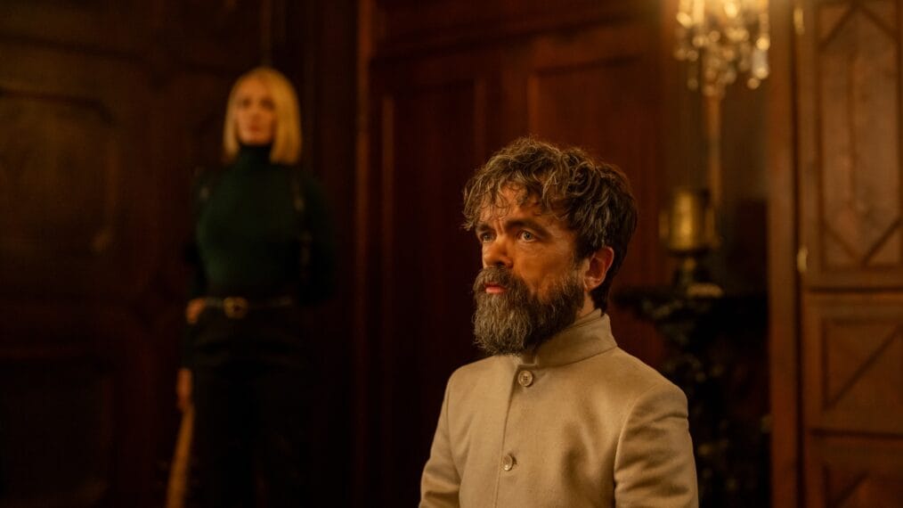 Peter Dinklage appears sitting in an elegant room, looking attentive with a woman standing behind him.