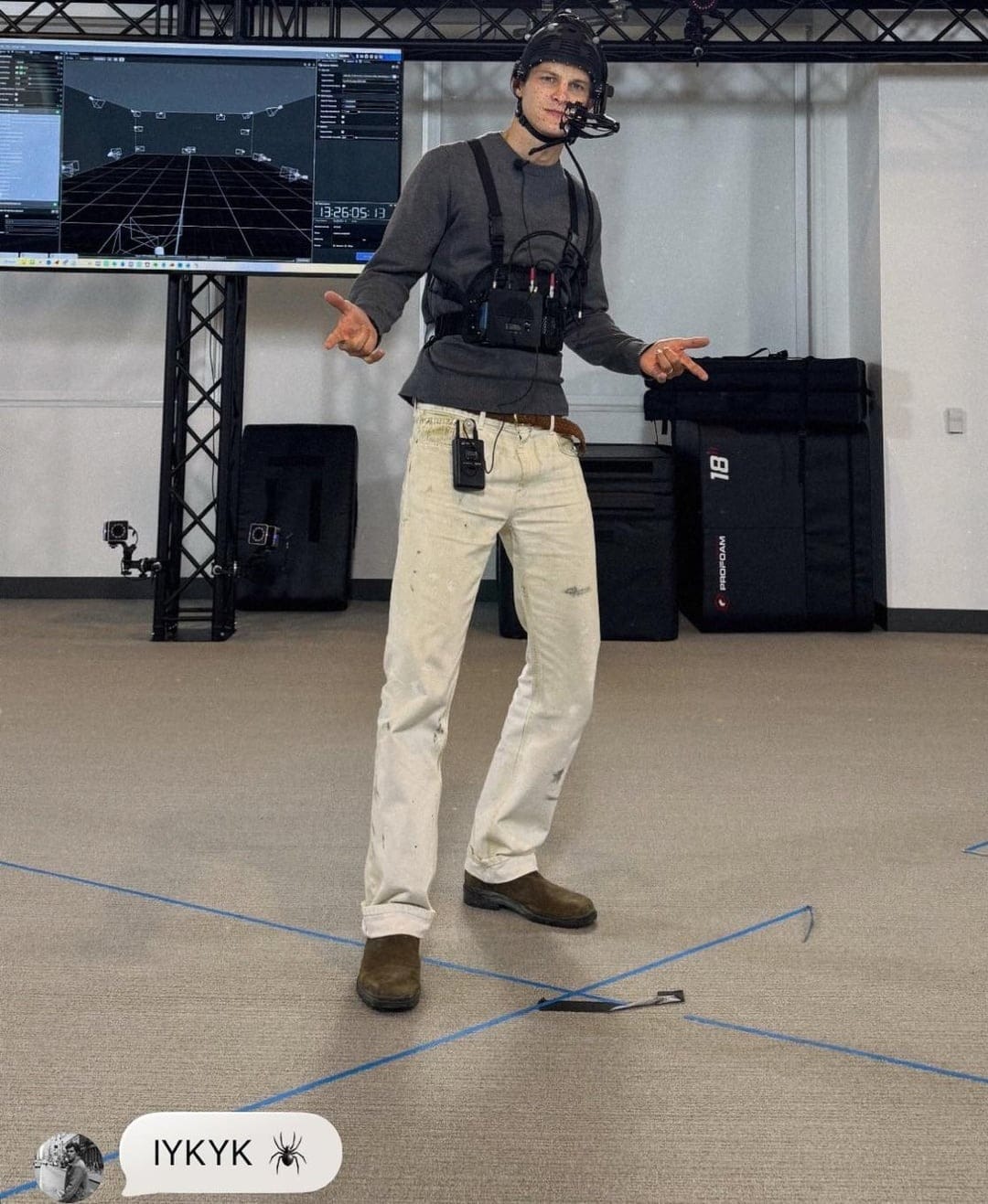 Person performing motion capture with sensors and a headset in a studio, with a digital display in the background.
