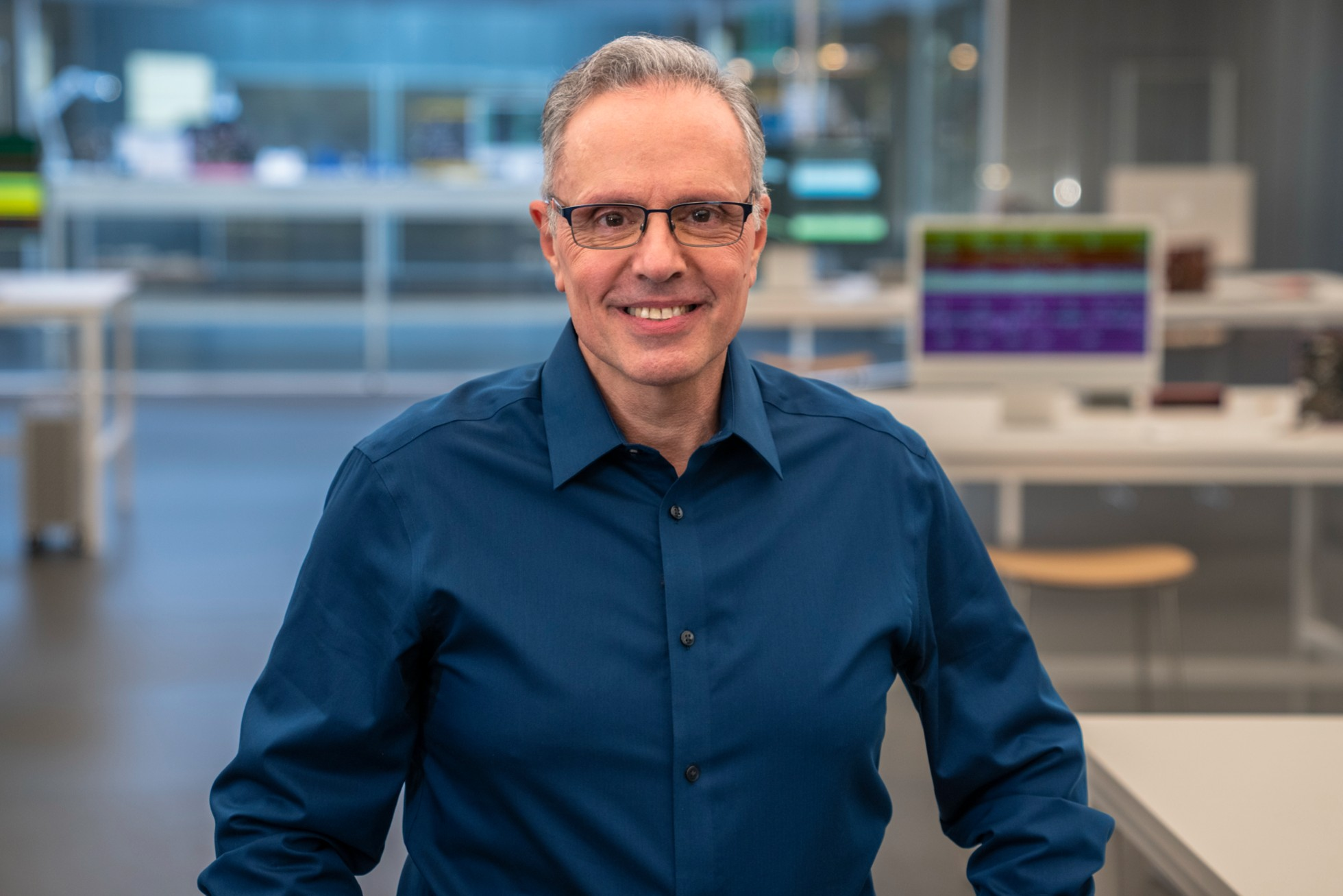 John Ternus smiling in a modern office environment with technology displays in the background.