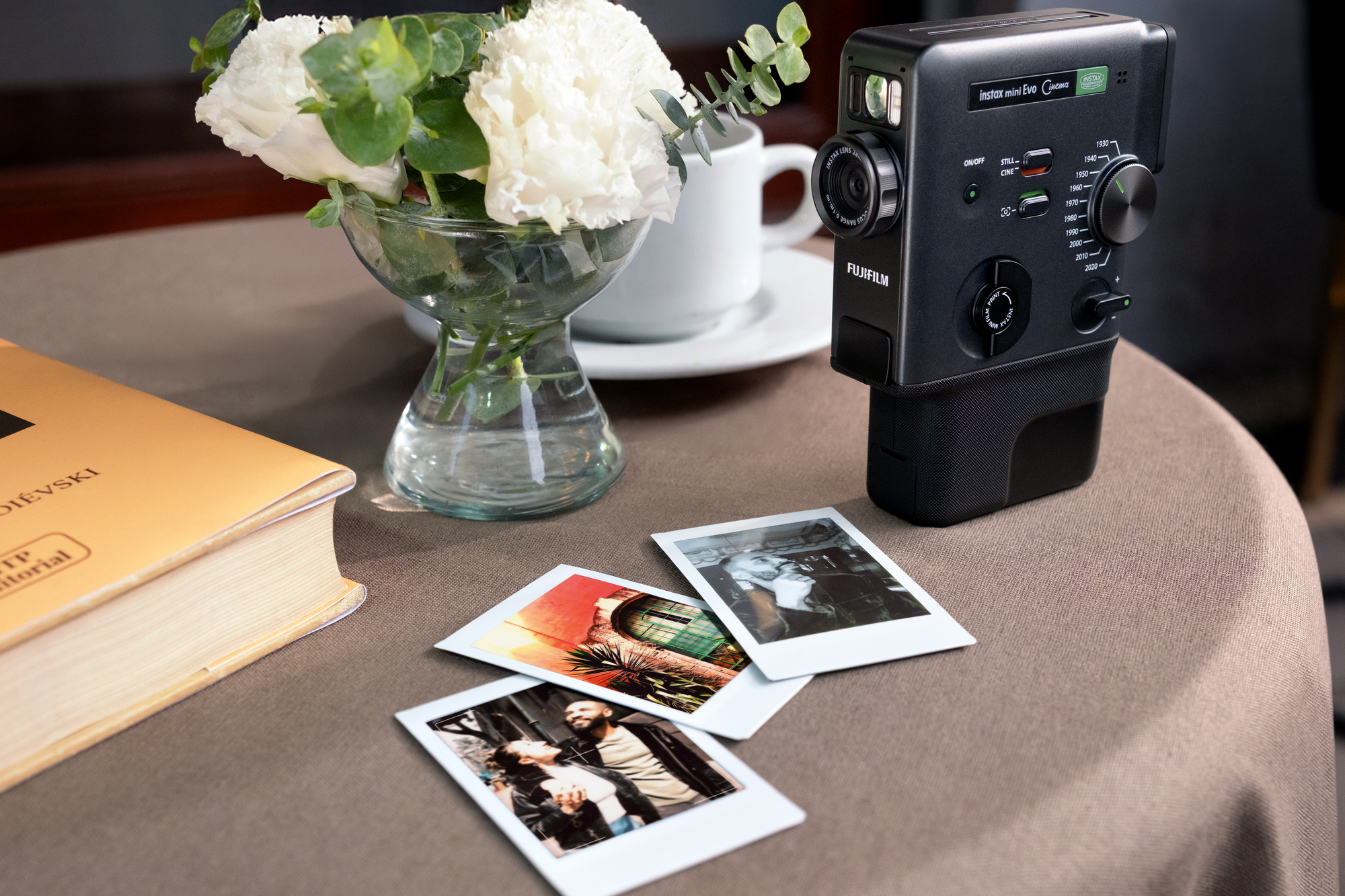 Fujifilm instax mini Evo Cinema camera beside instant prints on a table with a vase of flowers.