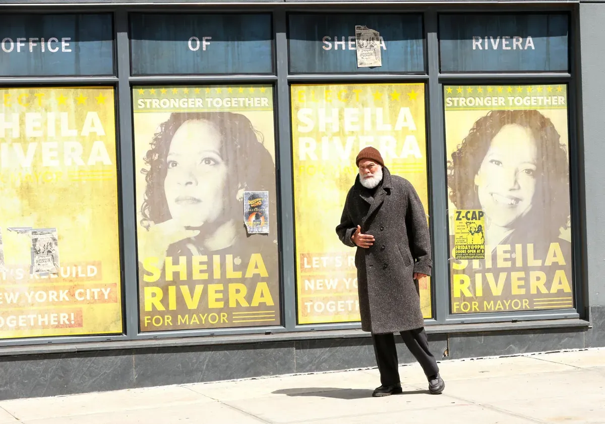 Kingpin stands in front of mayoral campaign posters for Sheila Rivera, dressed in a dark coat and hat.