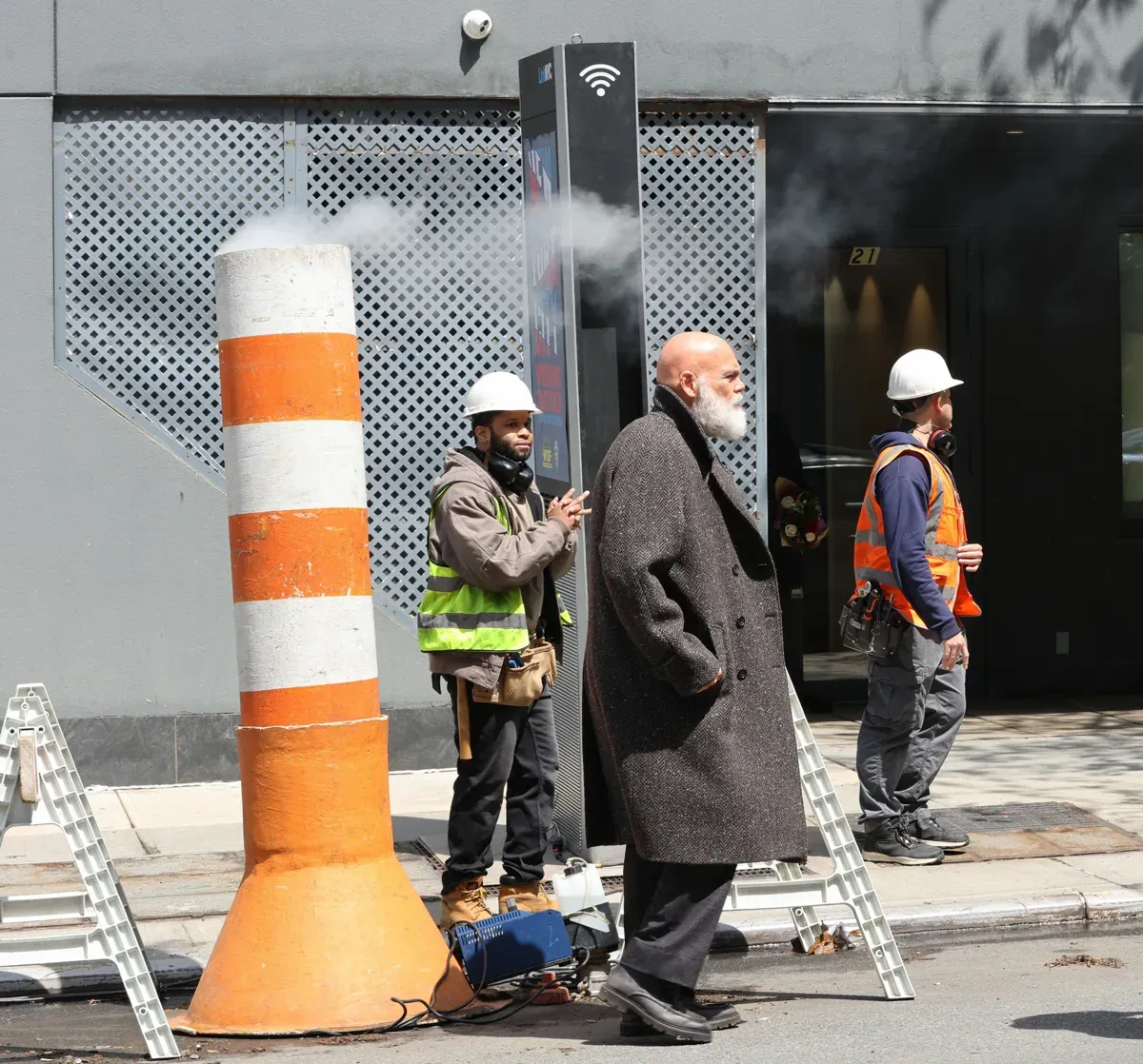 Kingpin casually stands by a striped construction barrier, watching as construction continues nearby.
