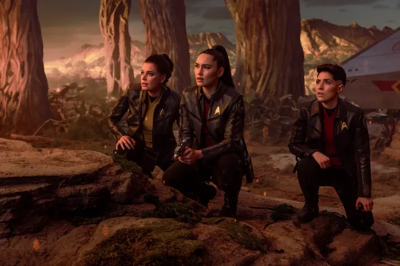 Three female characters in Starfleet jackets crouch in a forested alien environment.
