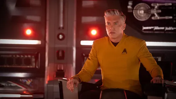 A character in a yellow Starfleet uniform sits in a command chair, looking alarmed.