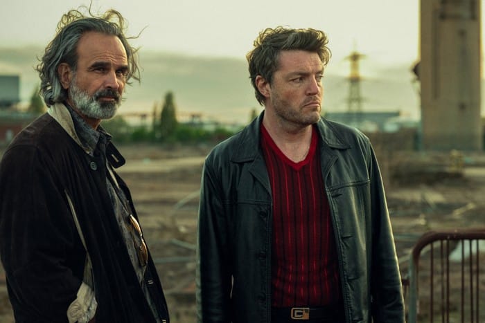 Two men in casual attire observe their surroundings in an abandoned area.