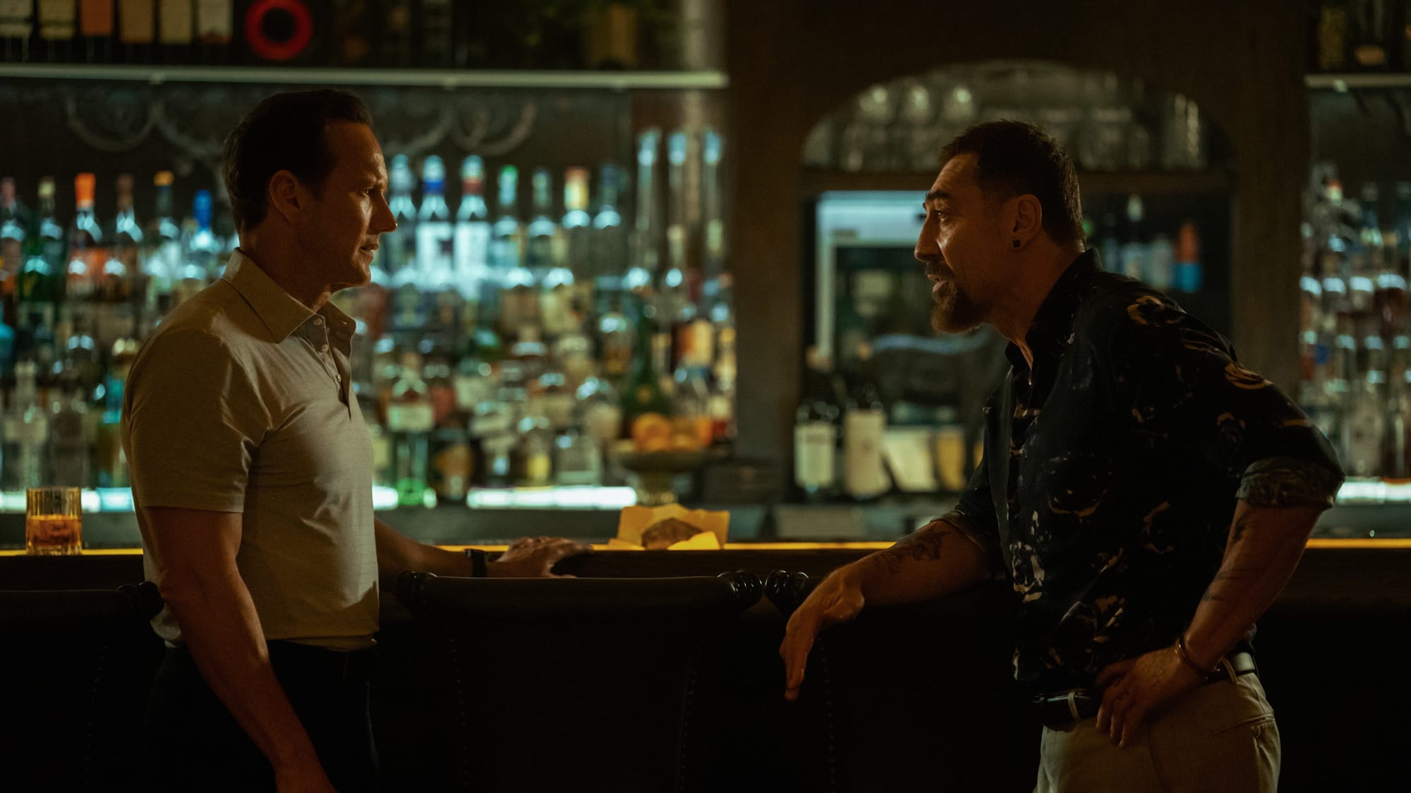 Two men conversing at a bar, surrounded by bottles, in a dimly lit establishment.