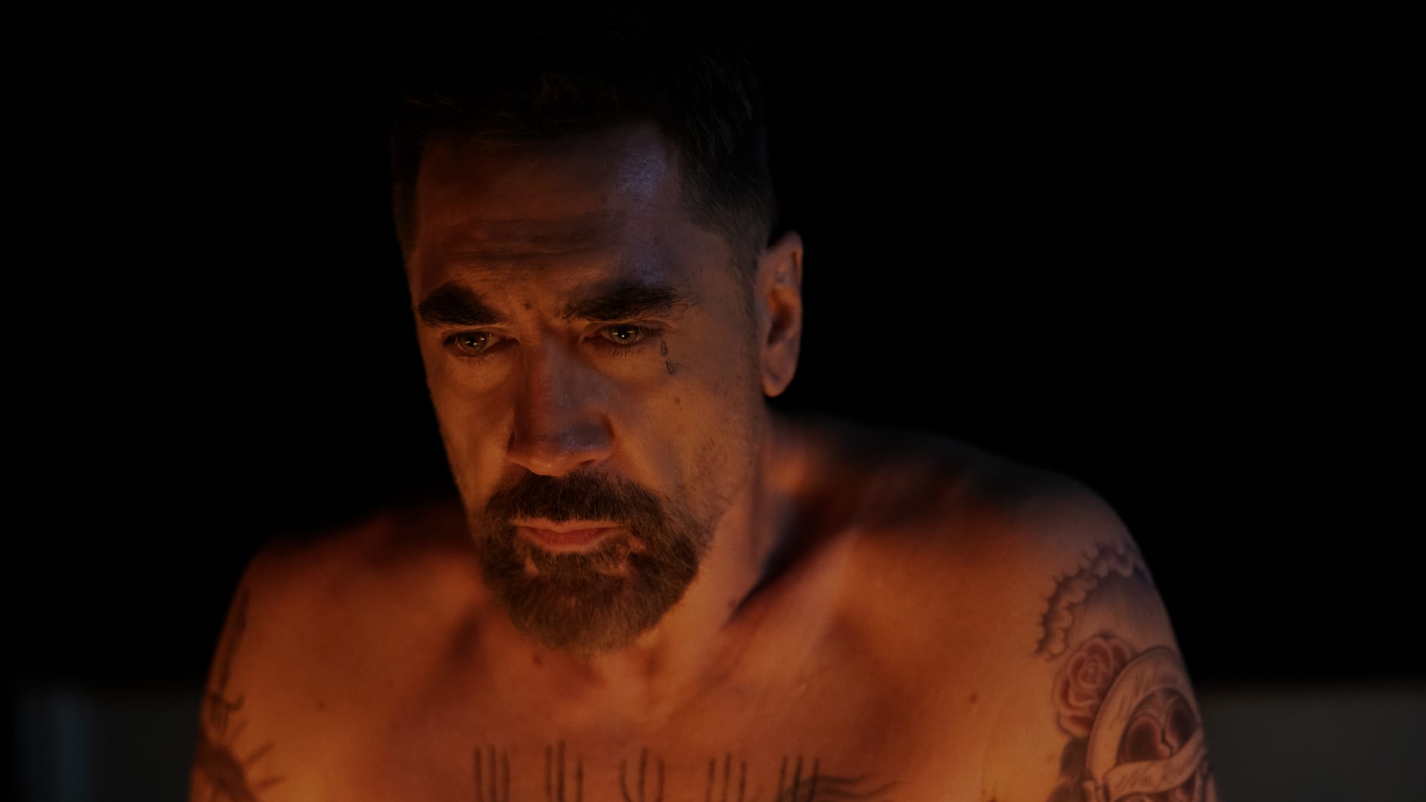 Close-up of a man with tattoos, looking thoughtfully at a flame in a dark setting.