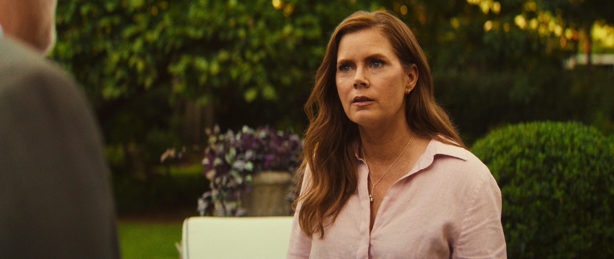 A woman with long hair wearing a pink shirt, speaking seriously in a garden setting.