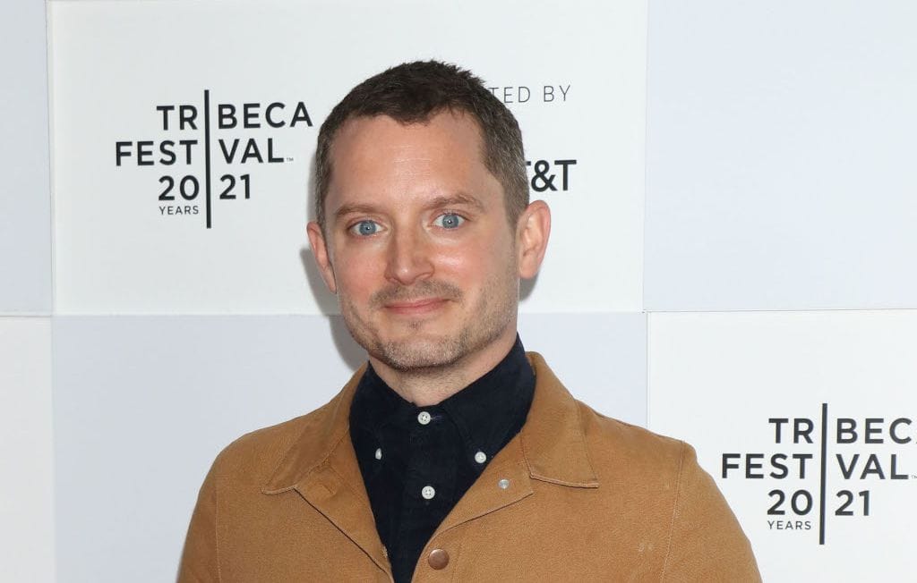 Elijah Wood smiling at the Tribeca Film Festival in a brown jacket.