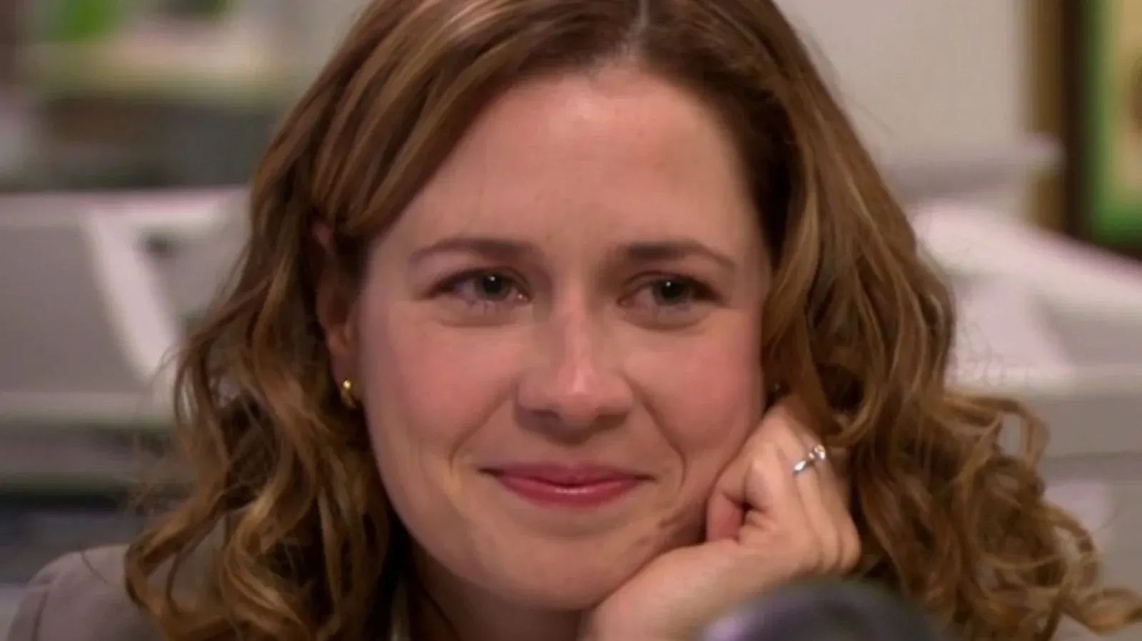 Pam Beesly smiling while resting her chin on her hand in an office setting.