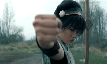 Toph, wearing a green outfit, clenches her fist while focusing intently in a natural setting.