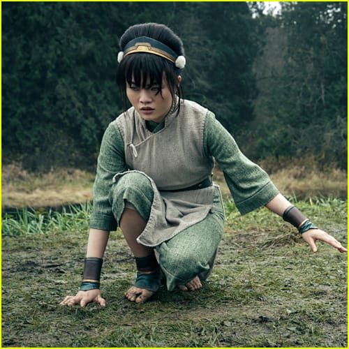 Toph crouches in a green outfit, preparing for action in a grassy environment.