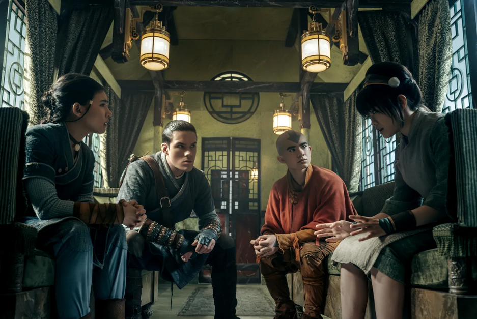 Four main characters sit together in a cozy, traditionally decorated room appearing deep in conversation.