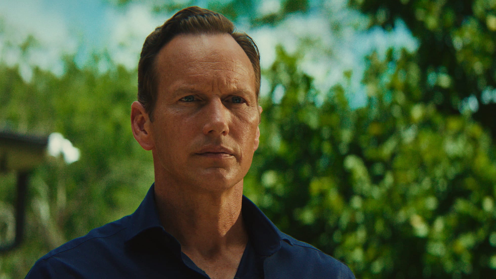 A close-up shot of a man with a serious expression, set against a lush background.