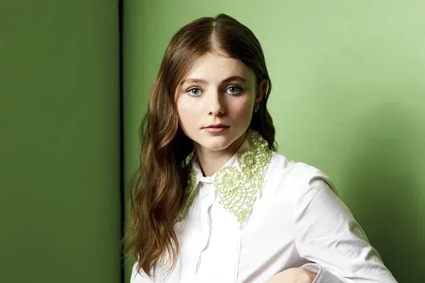Thomasin McKenzie poses against a green backdrop with stylish attire and a unique collar detail.