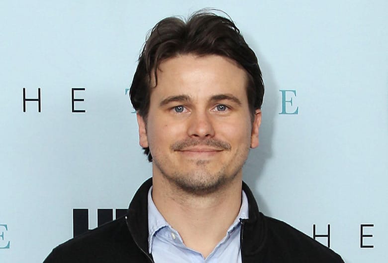 Jason Ritter poses for a photo at a promotional event, wearing a black jacket over a blue shirt.