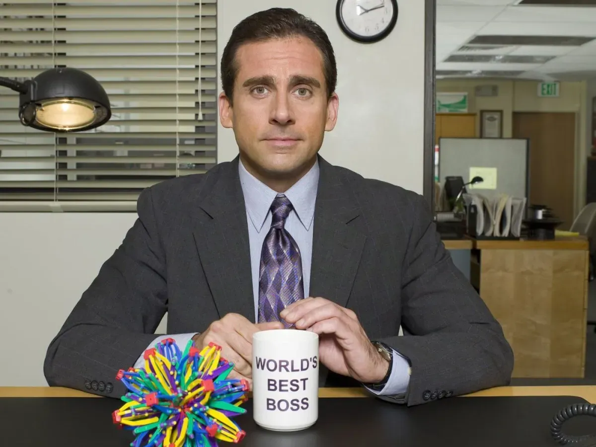 Steve Carell as Michael Scott, seated at a desk with a 'WORLD'S BEST BOS' mug and colorful desk toy.