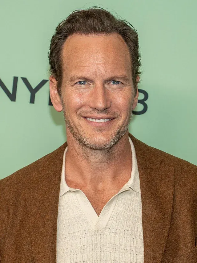 Patrick Wilson smiles at a promotional event, wearing a brown jacket over a light sweater.