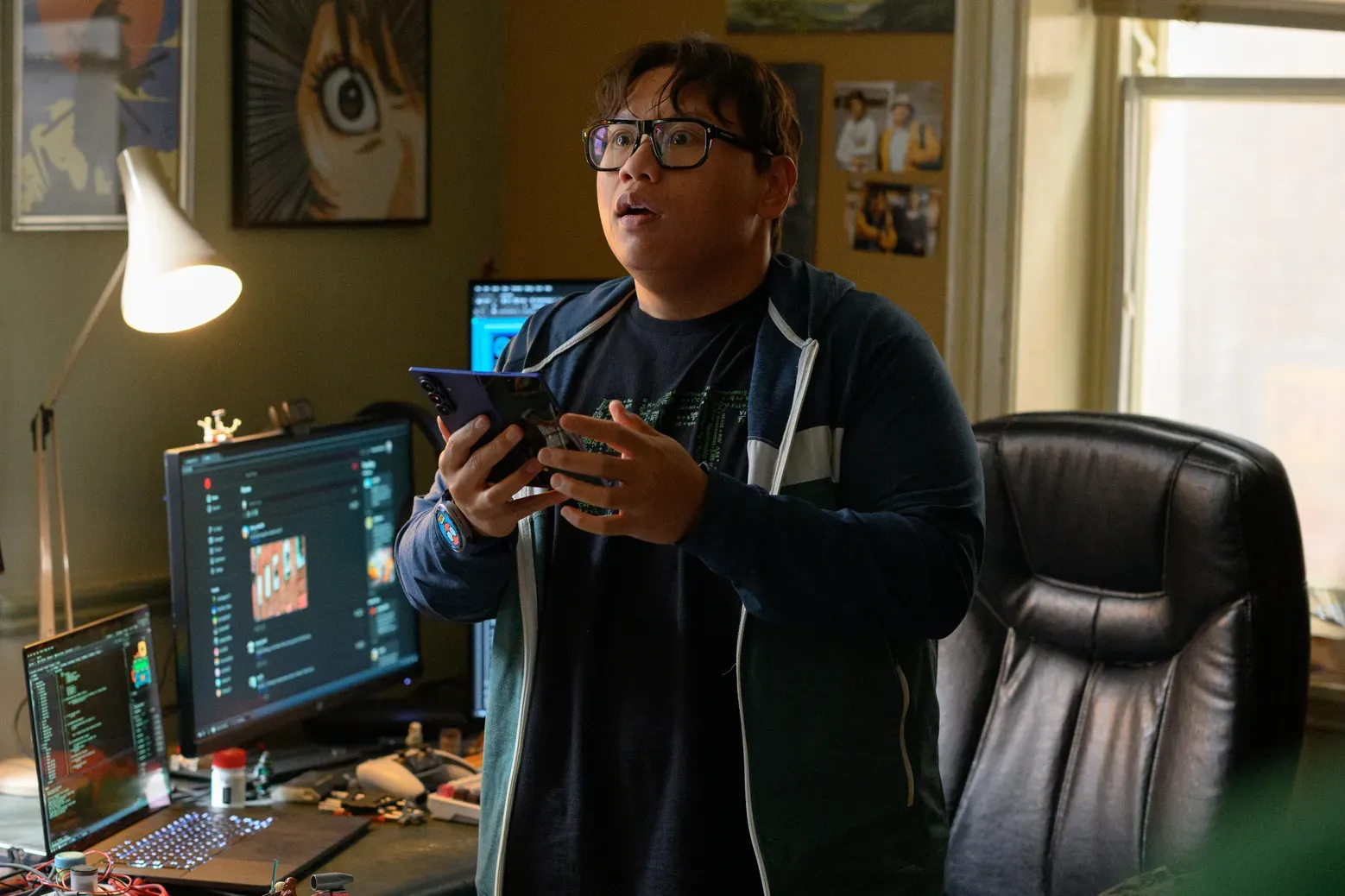 Ned Leeds holding a phone, looking surprised in a cluttered room with computer screens.