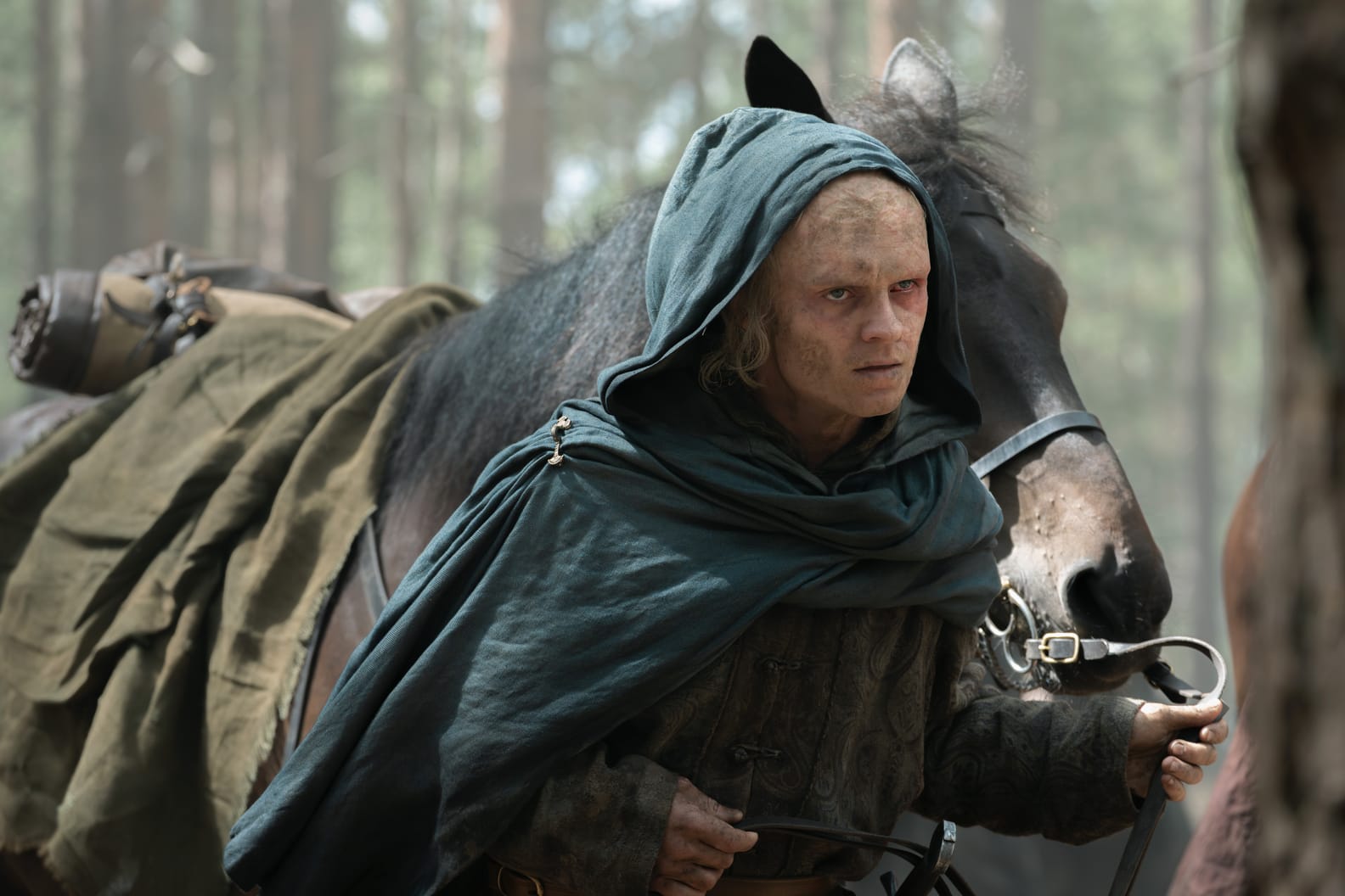 A rugged character leading a horse through a forest, dressed in tattered clothing.
