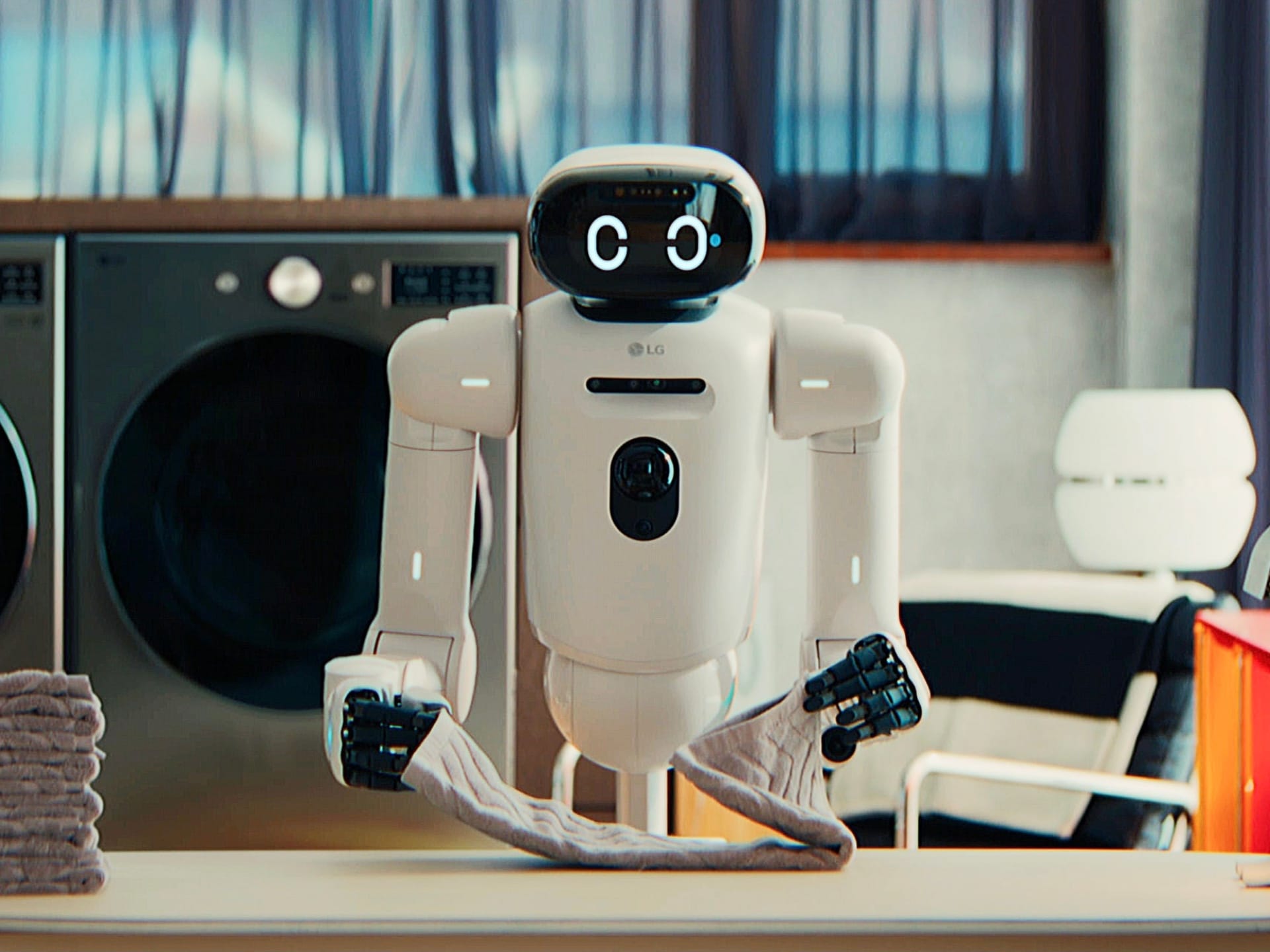 LG CLOiD is a white humanoid robot folding a gray towel on a table, with a stack of folded towels, a lamp, and washing machines in the background.