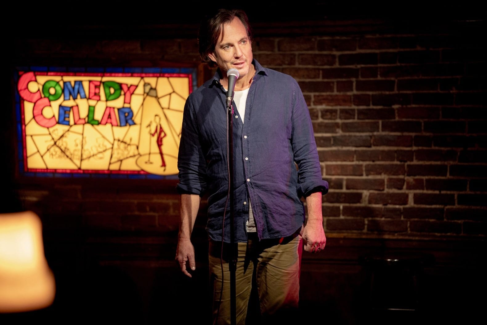 Stand-up comedian performing at a comedy club with colorful stained glass backdrop.
