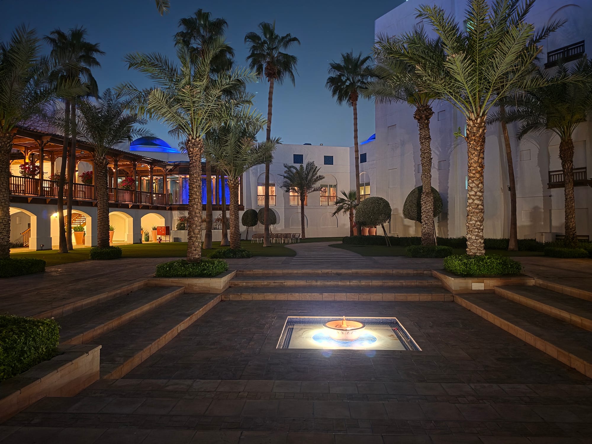 Night scene of a garden courtyard featuring a fountain and palm trees