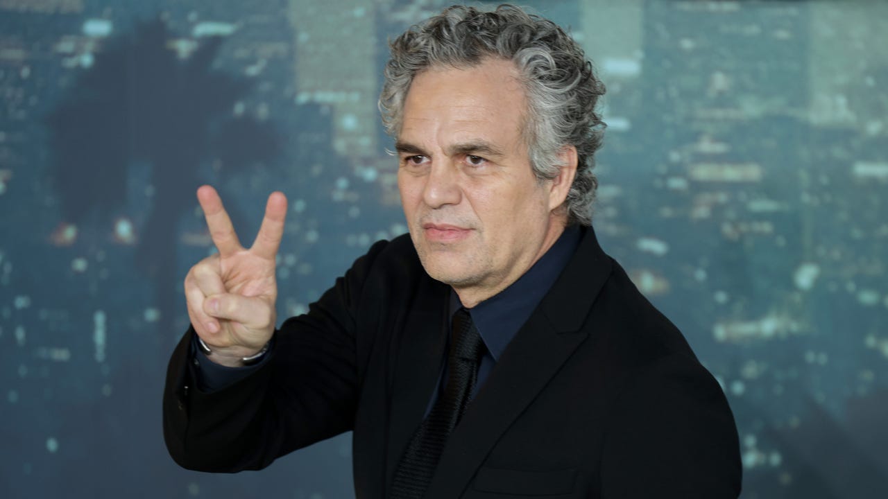 Mark Ruffalo in a dark suit raising two fingers in a peace sign at a red carpet event